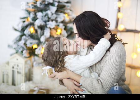 Schöne glückliche Mutter mit lher kleinen Tochter in Stricken Pullover sitzen auf dem Hintergrund von Weihnachten Innenraum umarmen zusammen und lächelnd Stockfoto