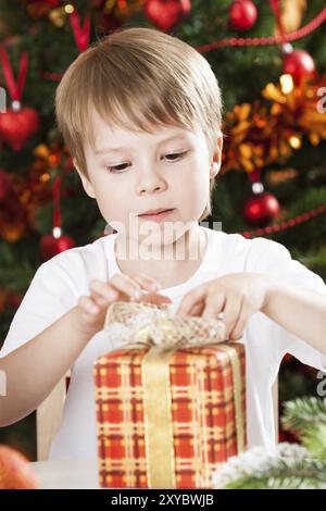 Überraschter Junge, der ein Geschenk vor dem Weihnachtsbaum mit Dekorationen öffnet Stockfoto