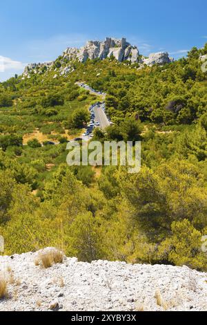 Von einer Klippe aus gesehen führt eine moderne Straße zu den Ruinen des Baux auf einem felsigen Berg in Les Baux de Provence, Frankreich, Europa Stockfoto