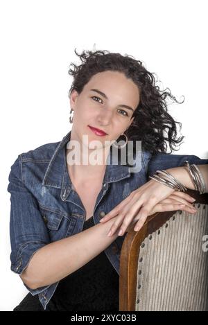 Ein junges Model, Mitte 20, sitzt auf einem hölzernen Stuhl mit einer blauen Jeansjacke Stockfoto