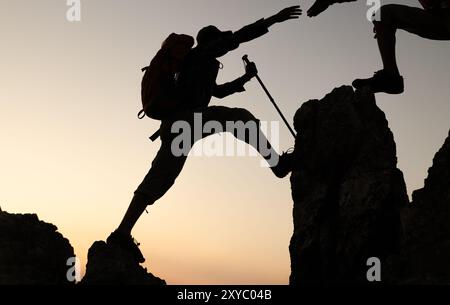 Silhouette eines Wanderers, der sich beim Sonnenuntergang gegenseitig beim Aufstieg auf einen Berg unterstützt. Menschen helfen und, Team arbeiten Konzept. Stockfoto