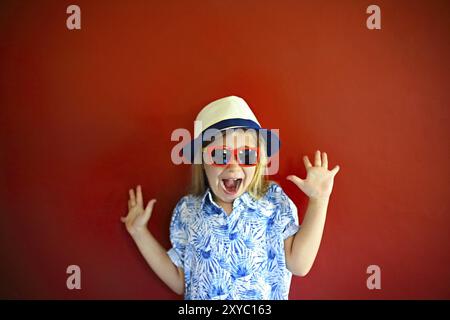 Ziemlich emothional Kind trägt einen Hut und eine Sonnenbrille auf einem roten Hintergrund. Platz kopieren, Tageslicht Stockfoto