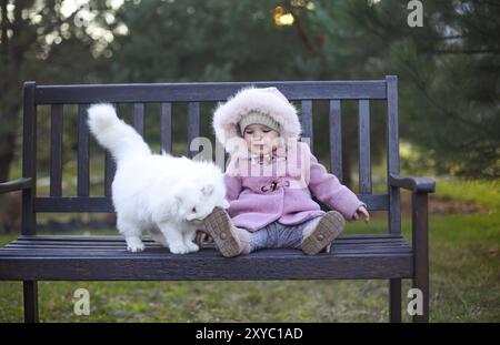 Kleinen niedlichen Baby Mädchen tragen Herbst Outfit und weiße Katze sitzt auf der Bank im Herbst Park Stockfoto