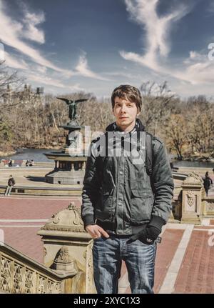 Ein junger, weißer, erwachsener Mann mit braunem Haar, der auf der Treppe im Central Park New york vor einem Brunnen steht, in Jacke und Schal überwintert Stockfoto