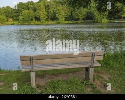 Eine Holzbank am Ufer eines Sees, umgeben von Gras, Bäumen und friedlicher Natur, Proebstingsee, Borken, Münsterland, Deutschland, Europa Stockfoto