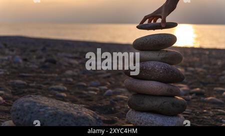 Ausgewogene Pyramide aus Kieselsteinen am Strand vor dem Hintergrund des Meeres bei Sonnenuntergang. Die Hand der Frau legt den letzten Stein in die Pyramide, den Kopierraum. Stockfoto