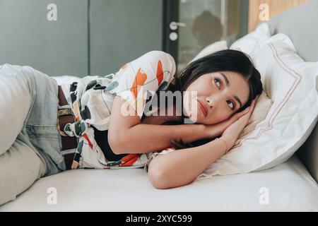 Ängstliche junge asiatische Frau mit hellem Denim und gemustertem Hemd liegt auf einem Bett und fühlt sich unwohl mit traurigem Ausdruck. Stockfoto