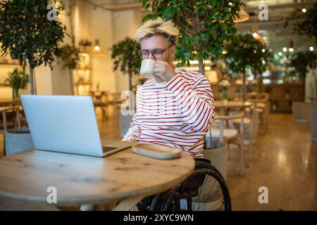 Interessierter Mann mit Behinderung trinkt Kaffee in einem Café, der freiberuflich online arbeitet und auf einem Laptop zusieht. Stockfoto