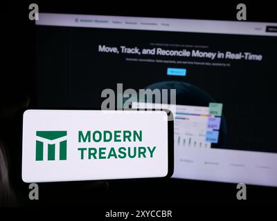 Person, die ein Mobiltelefon mit dem Logo der amerikanischen Geldbewegung Modern Treasury Corp. Vor der Webseite hält. Schwerpunkt auf dem Display des Telefons. Stockfoto