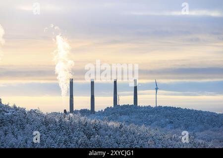 Vier Rauchstapel und ein Windkraftwerk bei einem Wald im Winter Stockfoto