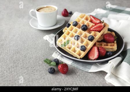 Süßes, köstliches Frühstück mit Waffeln, Erdbeeren und Heidelbeeren, Minzblättern und einer Tasse Kaffee auf betongrauem Hintergrund. Stockfoto