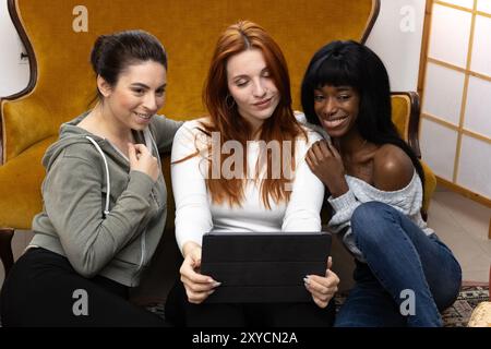 Drei verschiedene junge Frauen sitzen auf dem Boden in ihrer Wohnung, schauen sich ein Tablet an und machen ein Selfie Stockfoto
