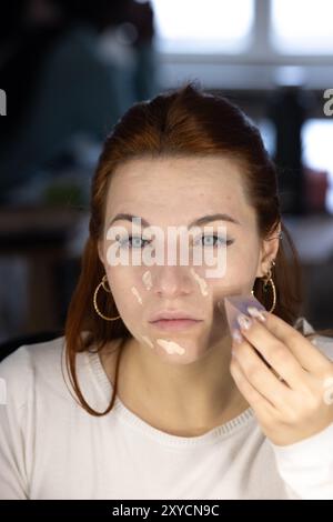 Die junge Frau trägt sorgfältig die Foundation auf ihr Gesicht auf und verwendet einen Schönheitsmixer für ein glattes und gleichmäßiges Finish Stockfoto