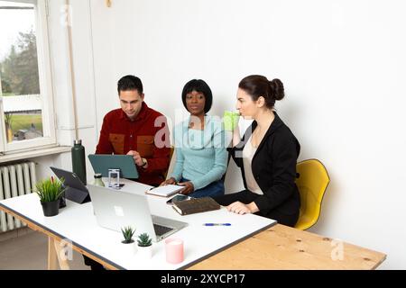 Drei junge Berufstätige sitzen an einem Schreibtisch, arbeiten gemeinsam an einem Projekt und nutzen Laptops und Tablets Stockfoto