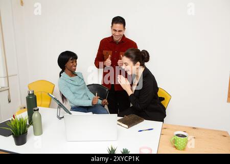 Drei glückliche und vielfältige Kollegen feiern im Büro einen Erfolg rund um einen Laptop Stockfoto