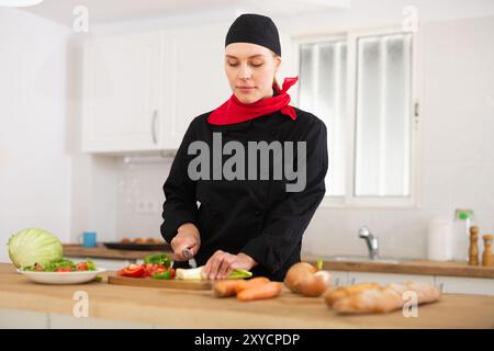 Positive weibliche Küchenchefin in schwarzer Uniform, die Gemüsegericht zubereitet Stockfoto