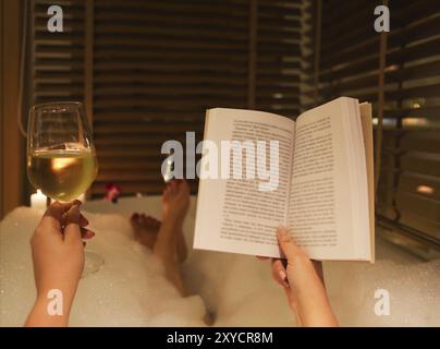 Zurück Blick auf die Füße der jungen Frau, das Buch zu lesen, und trinken Wein in der Badewanne mit Schaum und Kerzen Stockfoto