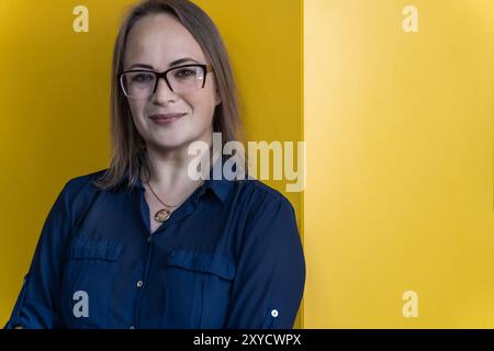 Lächelnde 40-jährige Frau in Brille mit blauem Hemd auf gelbem Hintergrund Stockfoto