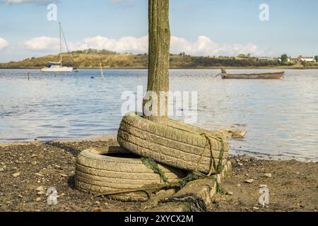 Alte Reifen und ein Holzpfosten in den Oare Marshes in der Nähe von Faversham, Kent, England, Großbritannien, mit Booten und der Isle of Sheppey im Hintergrund Stockfoto