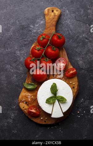 Ricotta-Käse mit Basilikum und Tomaten auf Schneidebrett aus Olivenholz auf schwarzem Schieferboden Stockfoto