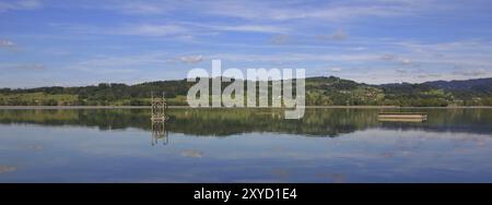 Grüne Hügel spiegeln sich im Lake Pfaeffikon. Frühlingsszene in der Schweiz. Badeplattform und Turm bereit für den Sommer Stockfoto