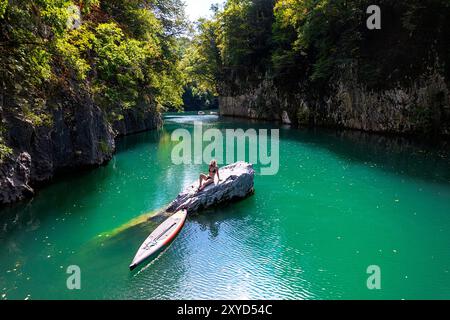 Touristikerin auf einer SUP-Reise, entspannend auf einem Felsen, Stand-up-Paddle-Boarder, die die Schlucht von Soca und den Fluss Idrijca in der Nähe von Most na Soci, Slowenien, erkunden Stockfoto