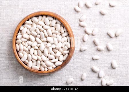 Weiße Bohnen in einer Holzschale auf Leinenstoff. Getrocknete kleine Samen, eine Sorte von Phaseolus vulgaris, die gewöhnliche Bohne, ein Mitglied der Familie der Hülsenfrüchte. Stockfoto