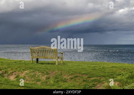 Eine Bank mit einem Regenbogen über der Nordsee Küste, im Benthall, Northumberland, England, UK gesehen Stockfoto