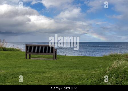 Eine Bank mit einem Regenbogen über der Nordsee Küste, im Benthall, Northumberland, England, UK gesehen Stockfoto