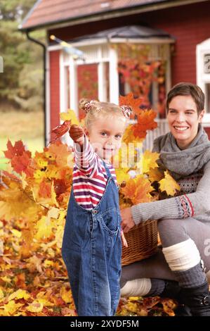 Fröhliches kleines Mädchen sammelt mit ihrer Mutter Herbstblätter in einem Korb. Im Hintergrund ein typisches rotes Holzhaus in Schweden. Lachende junge Mutter Stockfoto