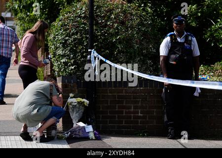 Ein Polizist steht neben einem Polizeikordel, während Mitglieder der Öffentlichkeit Blumenrabatte in der Overbury Street, nahe dem Tatort in der Rushmore Road, Clapton, im Osten Londons, ablegen, nachdem ein Mann in seinen 30ern erstochen wurde. Zwei Männer wurden verhaftet. Bilddatum: Donnerstag, 29. August 2024. Stockfoto