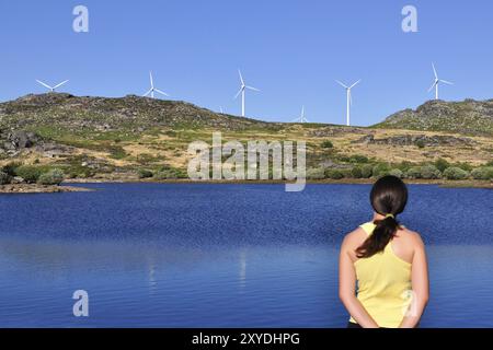 Frau, die auf eine Windmühle über einen See blickt. Der Fokus liegt auf den Windturbinen, und die Frau ist etwas außer Fokus Stockfoto