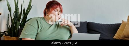 Eine große Frau mit rosa Haaren sitzt auf einer Couch in ihrem Haus und lächelt, während sie auf einen Laptop schaut. Stockfoto