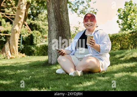 Eine Frau sitzt auf dem Gras in einem Park, lächelt und hält eine Tasse Kaffee. Stockfoto