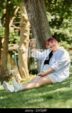 Eine Frau mit rosa Haaren sitzt auf dem Gras, lächelt und hält eine Kaffeetasse. Stockfoto