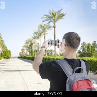 Ein junger Mann mit einem grau-roten Rucksack hinter den Schultern macht Fotos auf der Handygasse der Dattelpalmen Egypt Sharm El Sheikh South Shinai agai Stockfoto