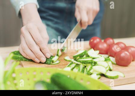 Nahaufnahme von weiblichen Händen, die in frisch geschnittene Gurken auf einem hölzernen Schneidebrett neben rosa Tomaten geschnitten wurden. Das Konzept der hausgemachten vegetarischen Küche und Stockfoto