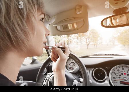 Niedliche blonde Frau mit kurzen Haaren, die Lippen färben, während sie am Steuer ihres Autos sitzt. Das Konzept, eine Frau zu fahren. Unangemessene Fahrstunden. D Stockfoto