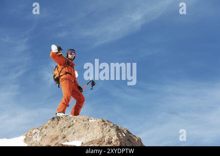 Porträt eines Skifahrers in einem orangen Overall mit einem Rucksack auf dem Rücken und Skiern auf den Schultern in einem Helm steht auf einem Felsen vor dem blauen Himmel. Kopieren Stockfoto