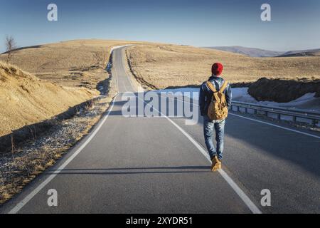 Ein bärtiger Hipster mit einem altmodischen Vintage-Rucksack, der Sonnenbrille mit rotem Hut und Jeansjacke trägt, und Jeans geht mit dem Rücken Stockfoto