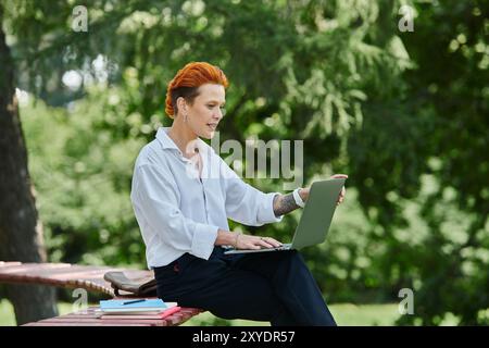 Eine Lehrerin mit rotem Haar sitzt auf einer Bank, vertieft in ihrem Laptop, umgeben von üppigem Grün. Stockfoto