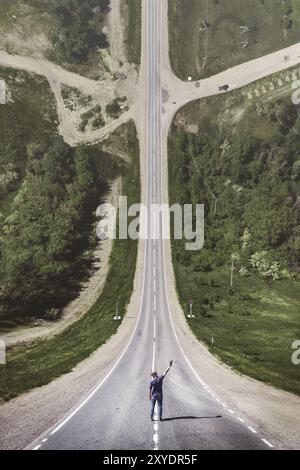 Ein junger Mann in einer Kappe mit einer Hand, die mit einer Konsole aus einer Drohne hochgezogen ist, steht auf einer Landstraße, an deren Ende sich ein Kreuz befindet Stockfoto