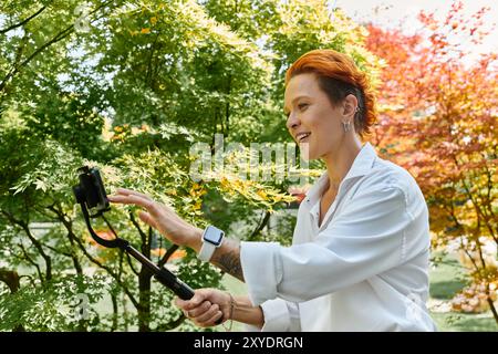 Eine Frau mit roten Haarfilmen mit einem Selfie-Stick in einer üppig grünen Campus-Umgebung. Stockfoto