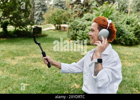 Eine Frau mit rotem Haar nimmt ein Video mit einem Selfie-Stick auf ihrem Handy auf und lächelt an einem sonnigen Tag Stockfoto