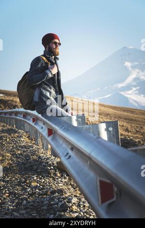 Bärtiger Touristen-Hipster-Mann in Sonnenbrille mit einem Rucksack, der auf einem Straßenrand sitzt und den Sonnenuntergang vor dem Hintergrund eines schneebedeckten M beobachtet Stockfoto