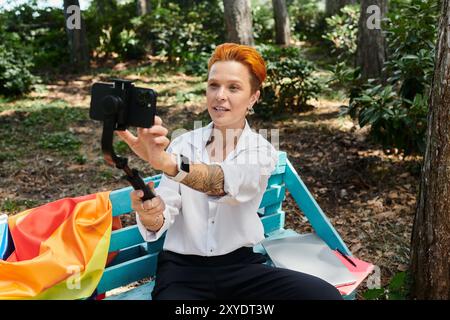Eine Frau mit orangefarbenen Haaren filmt sich auf einer blauen Bank mit einer Regenbogenfahne hinter sich. Stockfoto