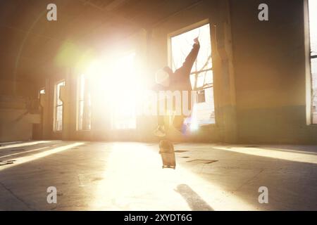 Ein junger Skater mit weißem Hut und schwarzem Sweatshirt macht einen Trick mit einem Skatesprung in einem verlassenen Gebäude im Hintergrund der untergehenden Sonne. Die Stockfoto