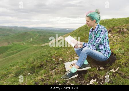 Porträt eines jungen Hipster-Mädchens mit mehrfarbigem Haar, das in den Bergen auf der Natur sitzt und ein Buch liest. Der Begriff des Lesens in der Natur. Bildungswesen Stockfoto