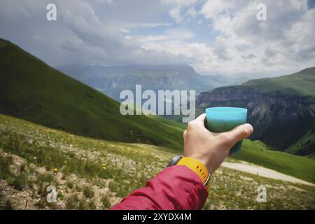 Aus erster Person betrachtet, wie die Hand eines Mannes eine Tasse Tee aus Plastik gegen ein Plateau aus grünen Hügeln hält und im Sommer einen wolkenlosen Himmel hat Stockfoto
