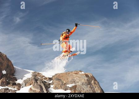 Ein Skirennläufer springt von hohen Felsen in den Bergen. Flusspulver aus dem Schnee hinter dem fliegenden Athleten Stockfoto